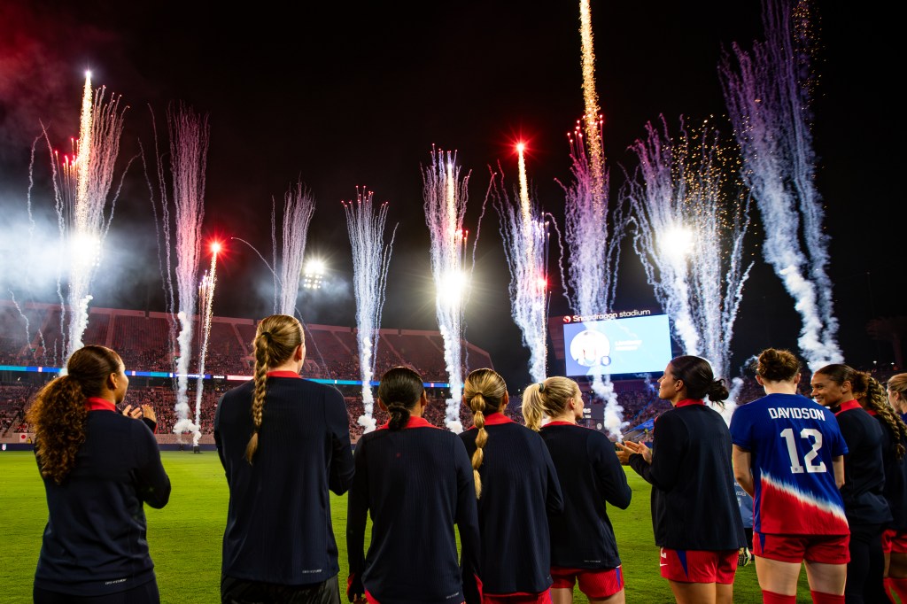 A group of female soccer players in uniforms stands in front of a stadium, celebrating with fireworks shooting into the night sky.