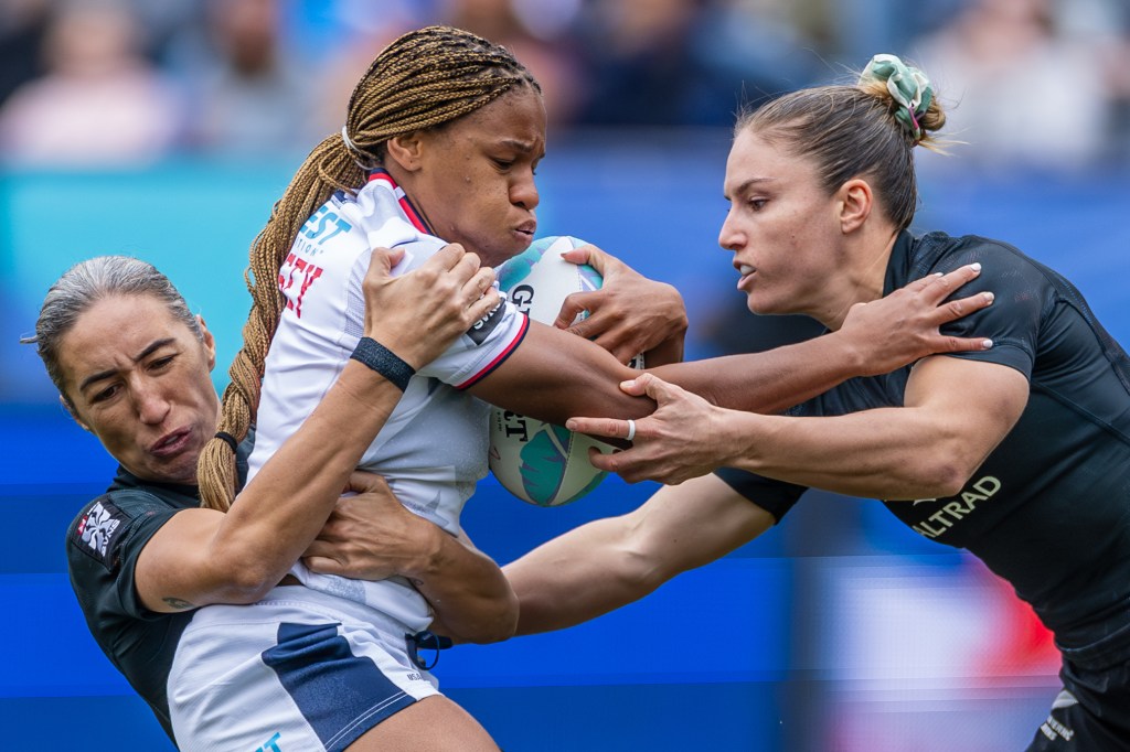 Two female rugby players engaged in a competitive tackle, with one player holding the rugby ball tightly while being pursued by opponents.