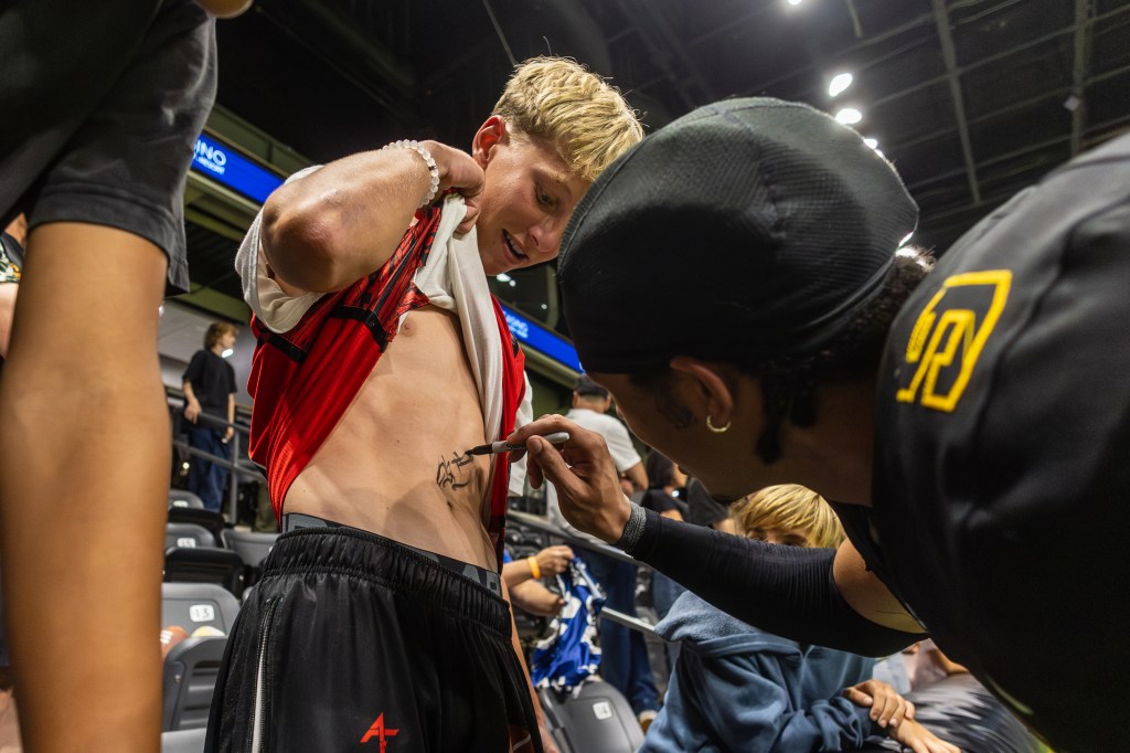 A young fan is getting a temporary tattoo applied to his side while wearing a jersey, with other attendees in the background at a sports event.