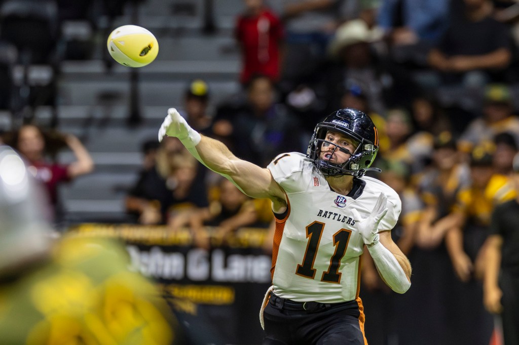 A player wearing a white jersey with 'Rattlers' printed on it, number 11, throwing a football during a game, with a blurred crowd in the background.